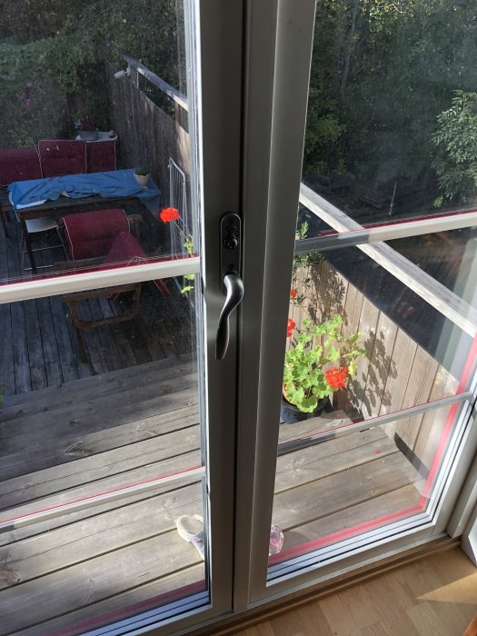 Vy genom ett stängt fönster som visar en dörrhandtag, låsmekanism och trädäck med möbler och blommor utanför.