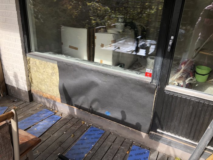 Halvfärdig installation av isolering och svart vindpapp på en altanvägg med omonterad panel och synligt isoleringsmaterial.