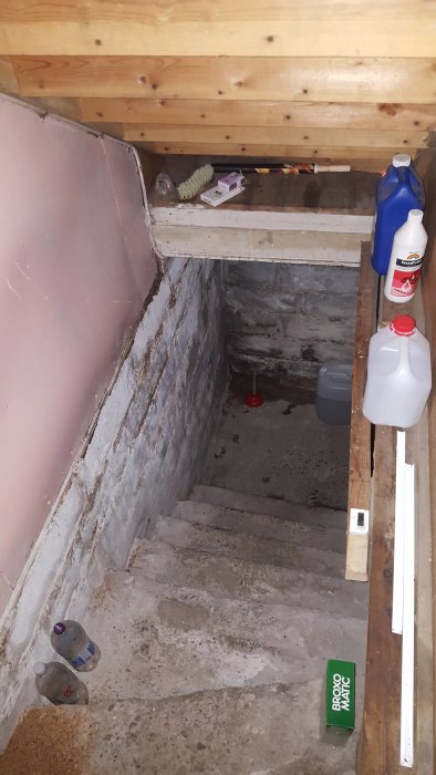 Sliten källartrappa i betong med stenväggar och viss bristfällig täckning av golvet, visar behov av renovering.