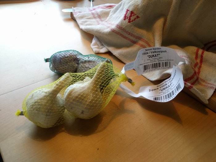 Nät med vitlöksklyftor 'Dukat' och 'Unikat' bredvid en kökshandduk på ett bord.