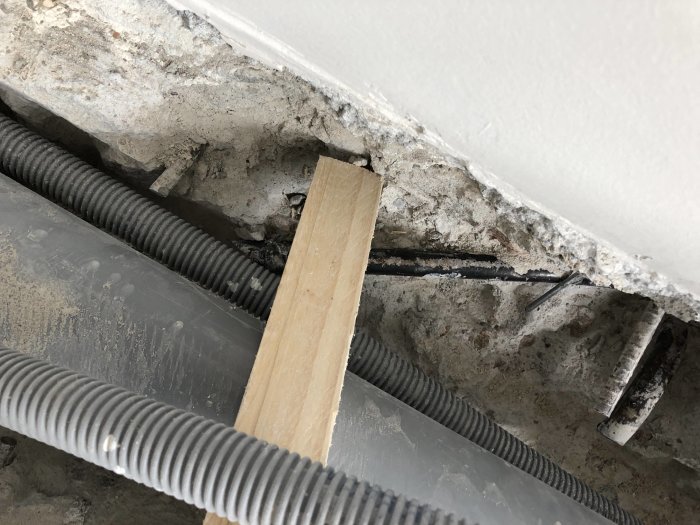 Uppbruten betong med pansarrör som visar ledningar som går in i väggen och ner i golvet, med en träplanka över.