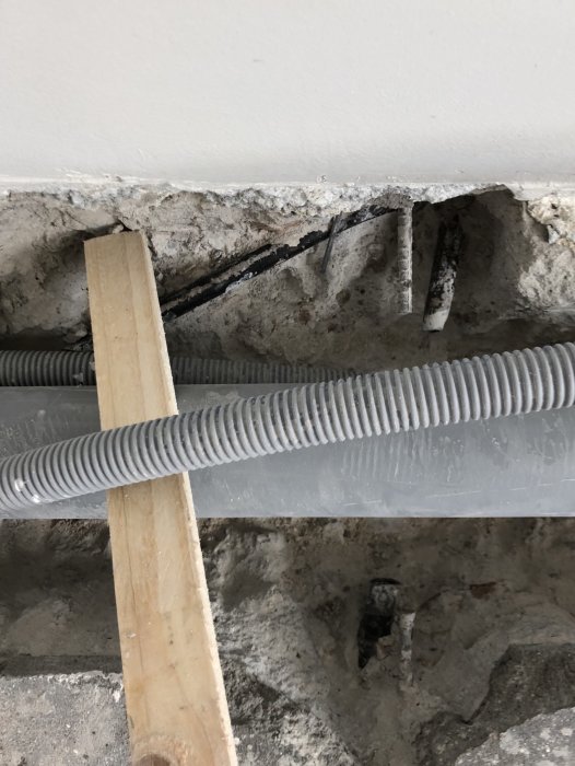 Uppbrutet betonggolv med exponerade pansarrör och synliga ledningar, stöttade av träreglar.