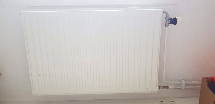 Vit vägghängd radiator med anslutningar och termostat i ett rum.