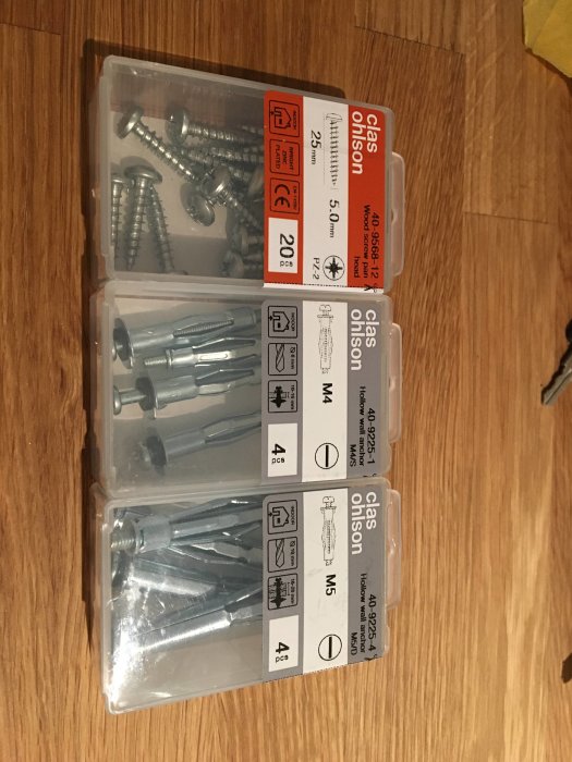 Förpackning med olika skruvar och pluggar på en träyta, Clas Ohlson-sortiment.
