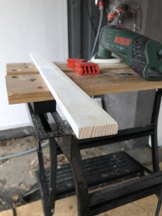 Nyslipad och målad träbit på arbetsbock med slippapper och sladdlös slipmaskin från Bosch i bakgrunden.