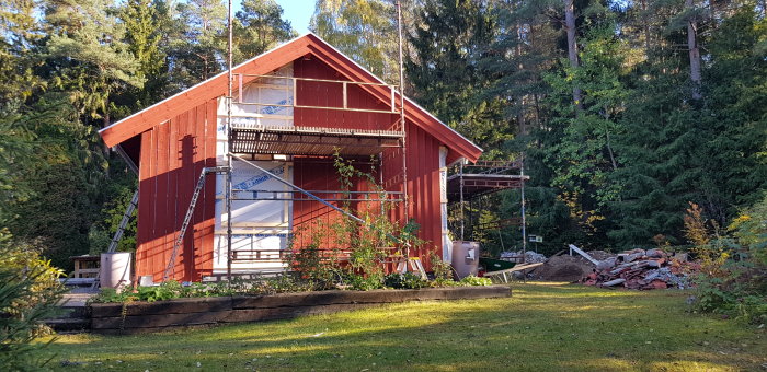 Rödfärgat hus under renovering med ställning och omonterade panelbitar, omgivet av träd och gräsmatta.