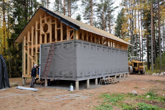 Hus under konstruktion klätt i vindväv med synlig stomme och person som arbetar.