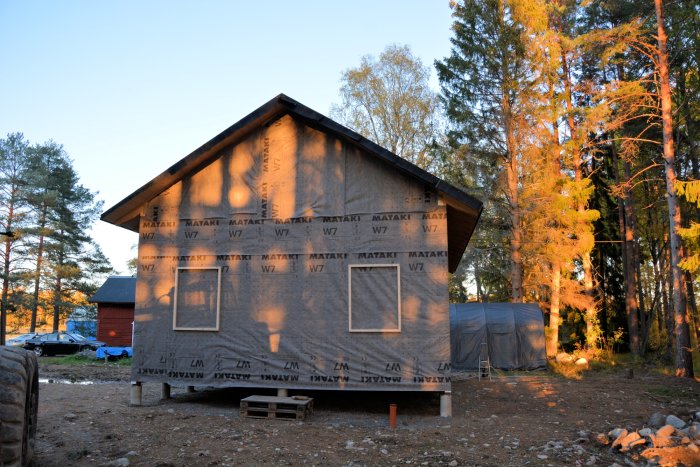 Hus under konstruktion klätt med vindväv och synliga 45-reglar kring fönsteröppning i skymningsljus.