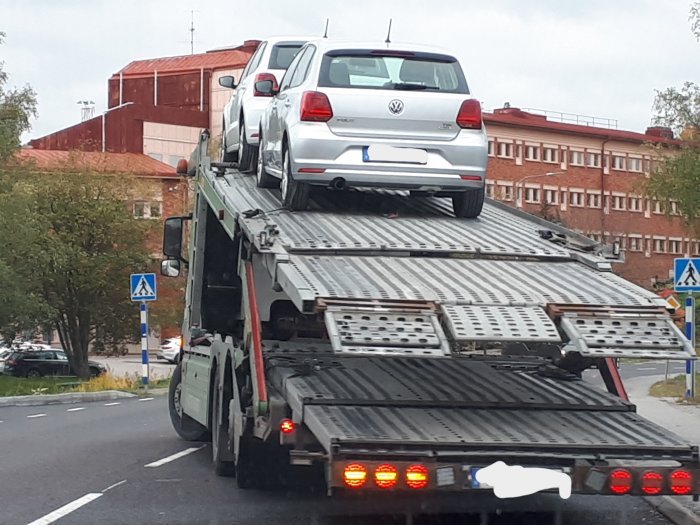 Vit Volkswagen bil på en biltransport längs en stadsgata med trafikskyltar och träd i bakgrunden.