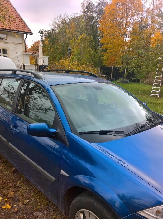 Blå Renault bil parkerad vid ett hus med höstlöv och en stege i bakgrunden.