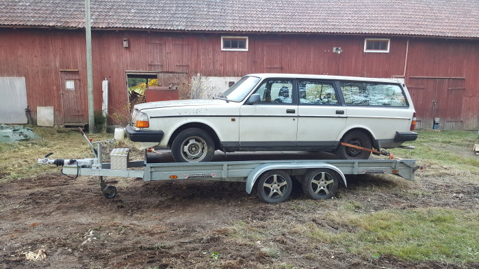 Vit äldre kombibil på släpvagn framför röd lada, säkrad för transport.