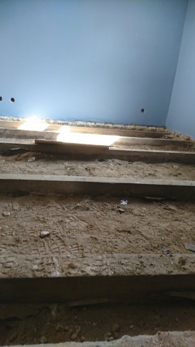 Upprivet golvexponerar träbalkar och sand, med en blåmålad vägg i bakgrunden, hemrenovering.