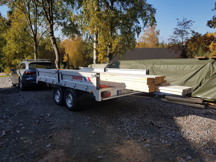Stort släp lastat med långa träreglar parkerat bakom en bil, redo för transport till byggprojekt.