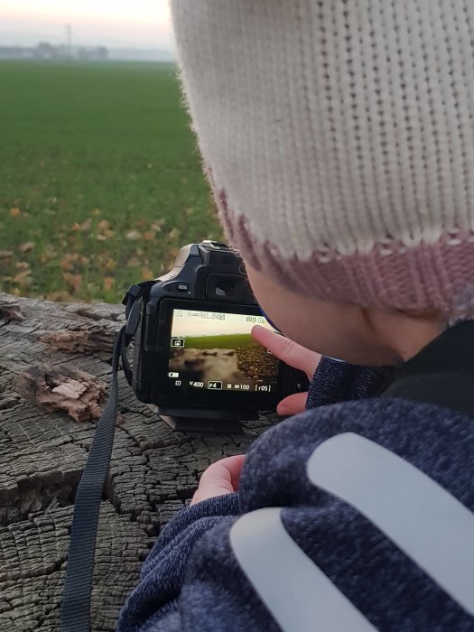 Barn i randig tröja tittar på en digitalkamera med oskarp bakgrund av ett grönt fält i skymningen.