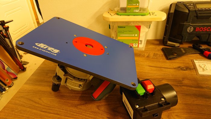 En överfräs med monterad kopierbas från Kreg på ett arbetsbord, omgiven av olika verktyg och väskor.