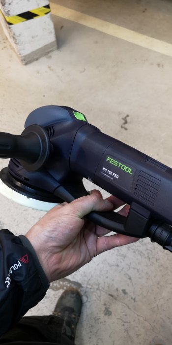 Hand håller en Festool RO 150 FEQ-slipsmaskin i en verkstadsmiljö.