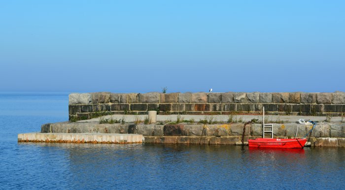 Klarblå himmel över lugnt hav med en röd båt vid stenbrygga, Simrishamn och Kivik.