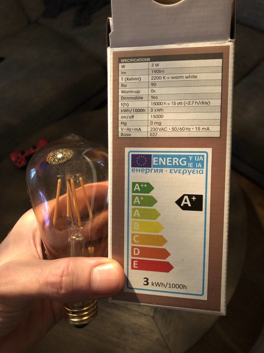 Hand håller en LED-lampa framför förpackning med energimärkning och tekniska specifikationer.