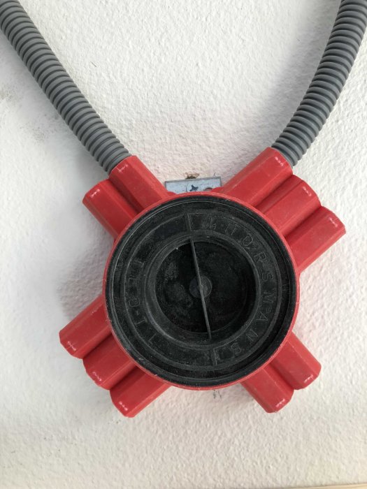 Röd ventilationsfläns med ansluten grå böjlig slang mot vit vägg.