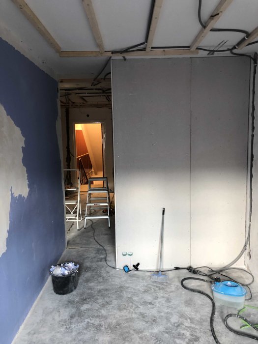 Renoveringsarbete med oslipade gipsskivor på vägg och målning påbörjad, byggmaterial och verktyg på betonggolv.