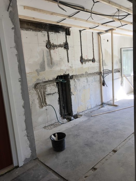 Renoveringsarbete på vägg med el- och vvs-installationer och en svart hink på betonggolv.