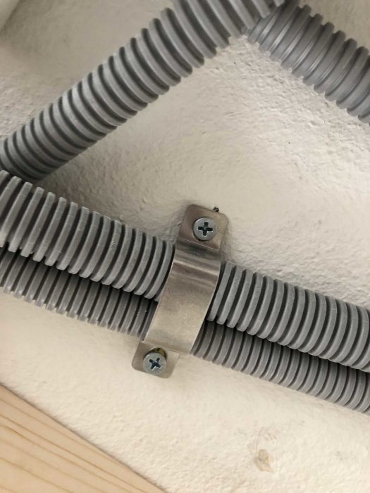 Korrugerad kabelskyddsslang fastsatt i hörnet av ett rum med metallklämma och skruvar.