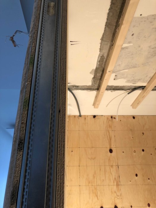 Del av ett byggnadsstomme med en metallprofilskena, oskyddade elledningar och en plywoodvägg.