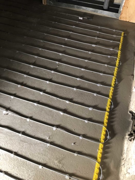 Nyinstallerat värmegolv med klammer och guldmeter på betongunderlag.