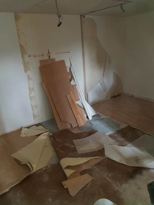 Renoveringsarbete i rum med borttaget golv, trasiga väggfragment och upprivna bruna mattbitar på betonggolvet.