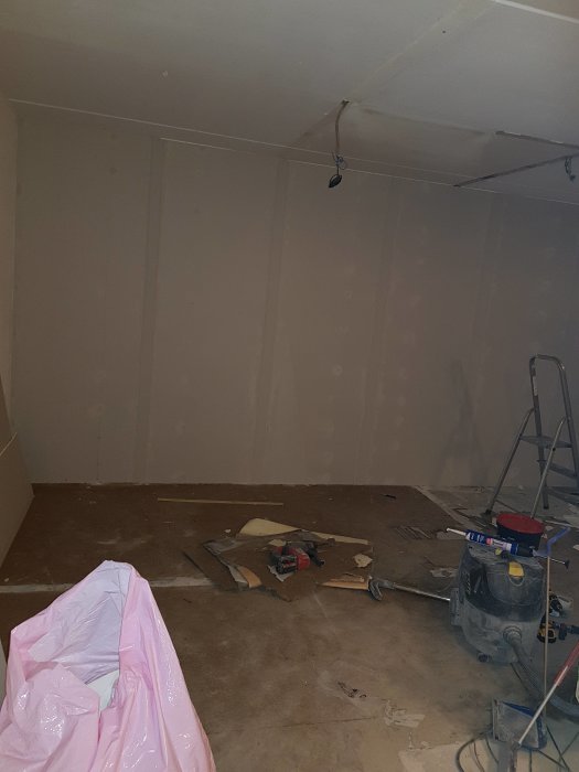 Renoveringsarbete i ett kök med oslipat betonggolv och nyuppsatta gipsskivor på väggen, ombyggnad pågår.