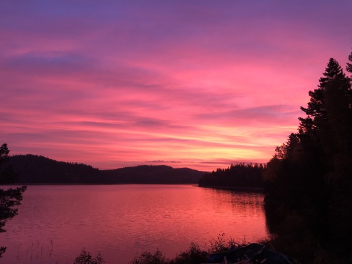 Vibrant pink and purple sunrise over a calm lake with silhouette of trees and hills.