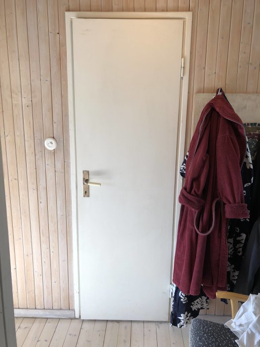 Vit innerdörr med fals monterad i trävägg, klädhängare med röd jacka till höger.