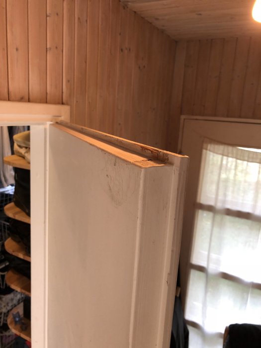Vit dörrkant med fals mot trävägg i rum, synlig skada och slitage på dörrens övre hörn.