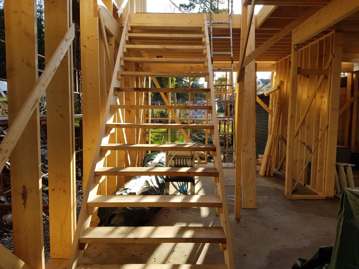 Träkonstruktion av en byggnad med tre klara väggar, tillfällig trätrappa och en pelare i hörnet.
