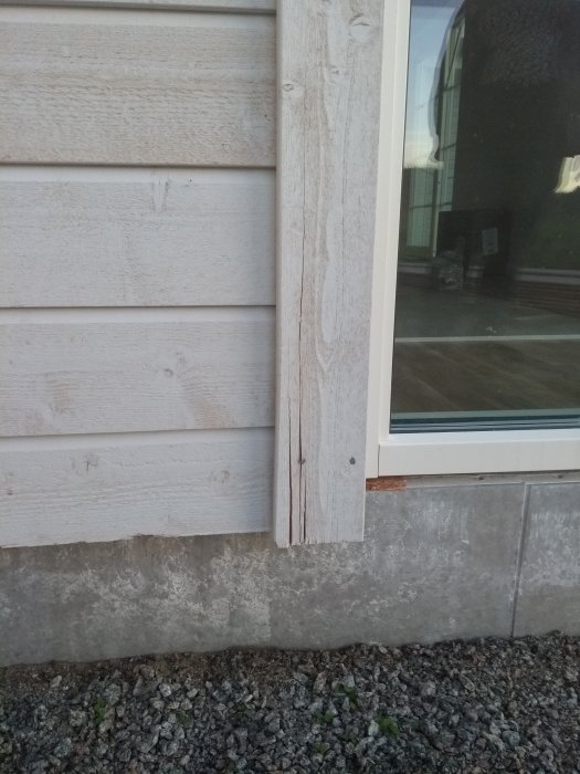 Synlig spricka i en vertikal träbräda vid husets hörn ovanför en betongsockel.
