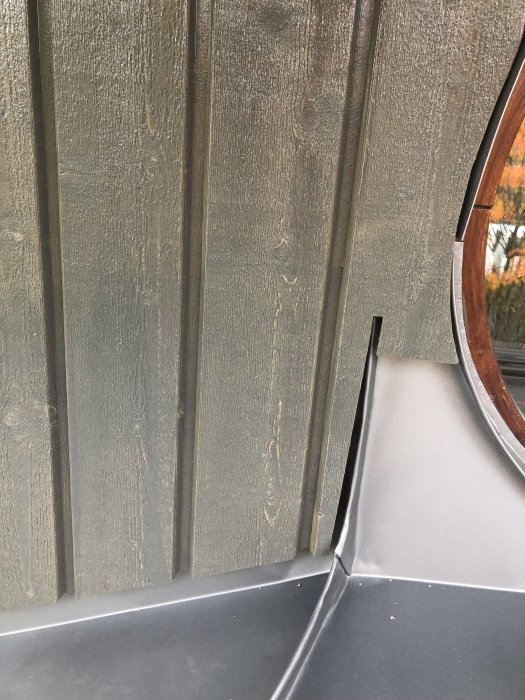 Vertikal träpanel nära entré med silvergrå plåtdetaljer och en del av ett rundat fönster.