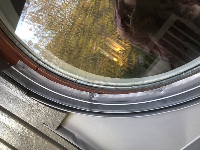 Nyinstallerat silver/grått överbleck på fönster, med träpanel och reflektion av träd i glaset.