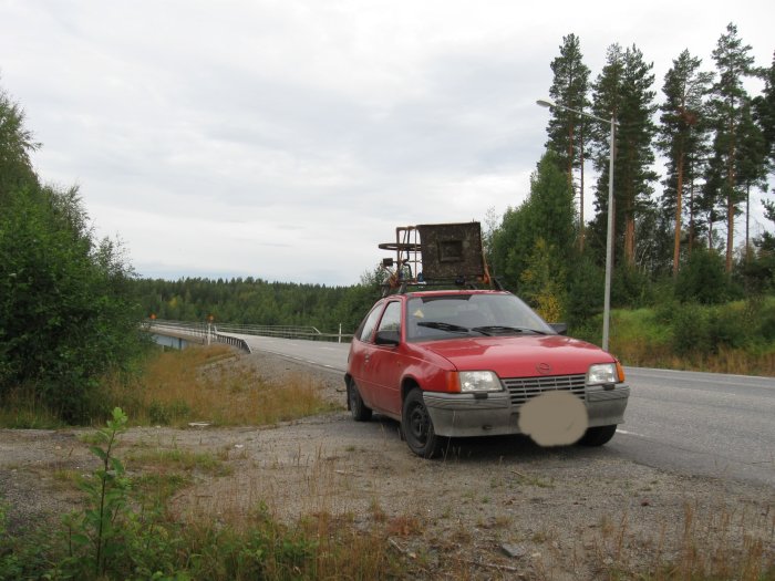 Röd Opel Kadett E från 1984 parkerad vid vägkanten med ett fynd på takräcket, i ett landskap med träd och gräs.