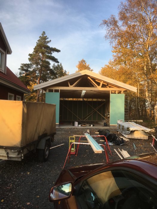 Pågående bygge av ett garage med uppförda väggar och takstomme, material och verktyg utspridda framför.