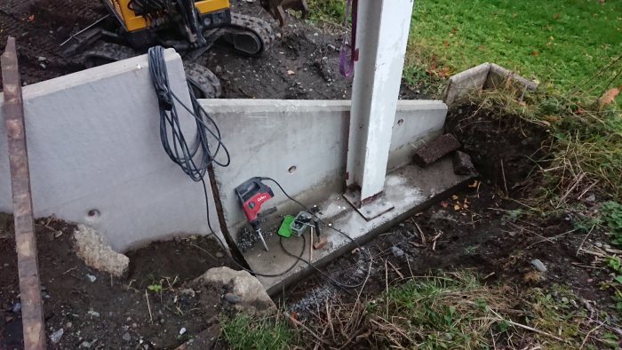 Betongfundament med stålpelare, borr och kablar, omgivet av grävd jord och gräsmatta.