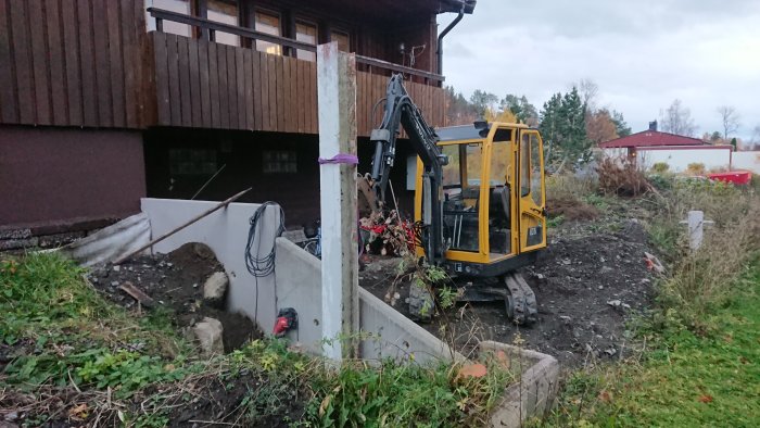 Grävmaskin nära hus med ojämna L-stöd och pelararbeten för balkong/carportprojekt.