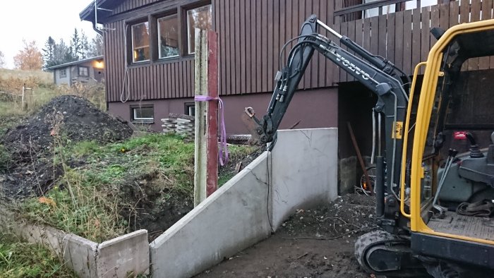 Grävare vid byggplats med jordhög, L-stöd och pelare vid hus i arbete med carport/balkong.