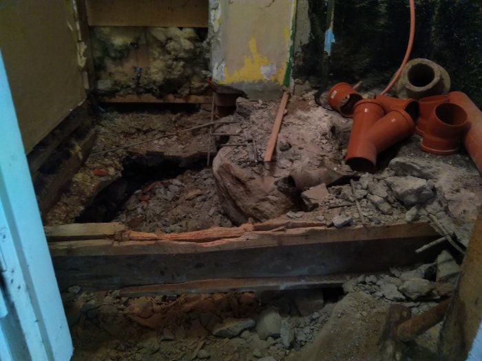 Rörlagda trasiga betonggolv med en stor sten, murat material och träbjälkar vid en murstock i renovering.