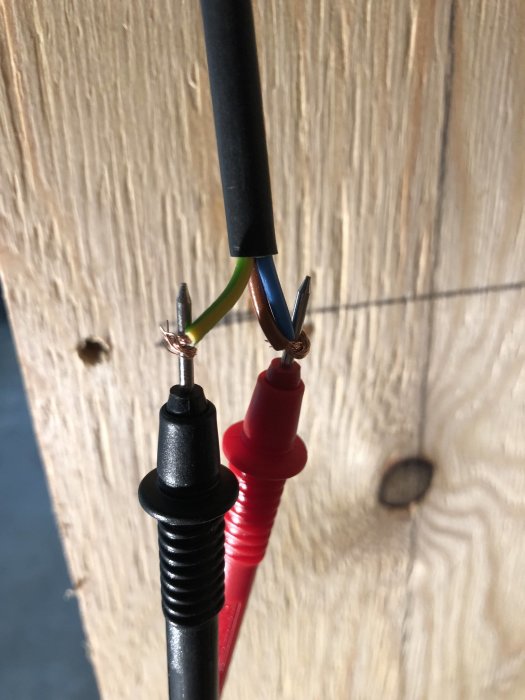 Elkabel med avskalade ändar anslutna till svart och rött mätinstrument mot träbakgrund.