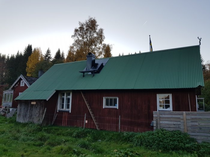 Röd stuga med grönt tak där skorstenen och luckan är klädda i plåt, omgiven av natur och staket.