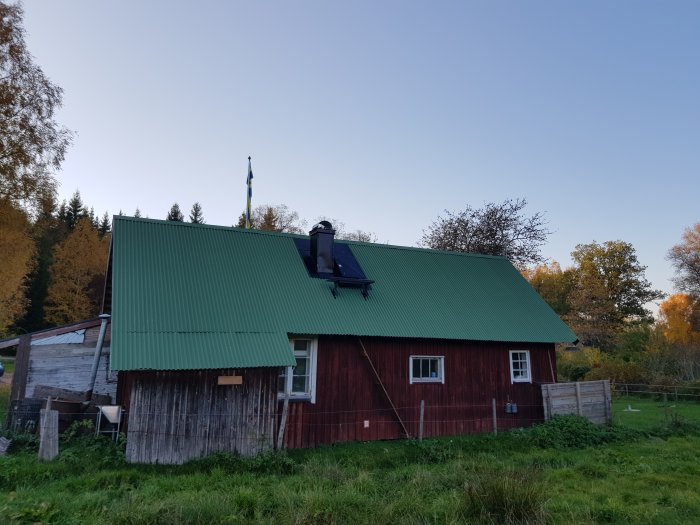 Röd träbyggnad med nyklätt skorsten i plåt, omgiven av gröna ängar och höstträd under klar himmel.