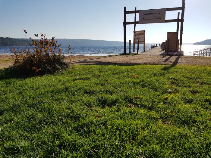 En solig strand med gräsmatta i förgrunden och välkomstskylt till "Stilla Sand" med bakgrund av lugnt vatten.