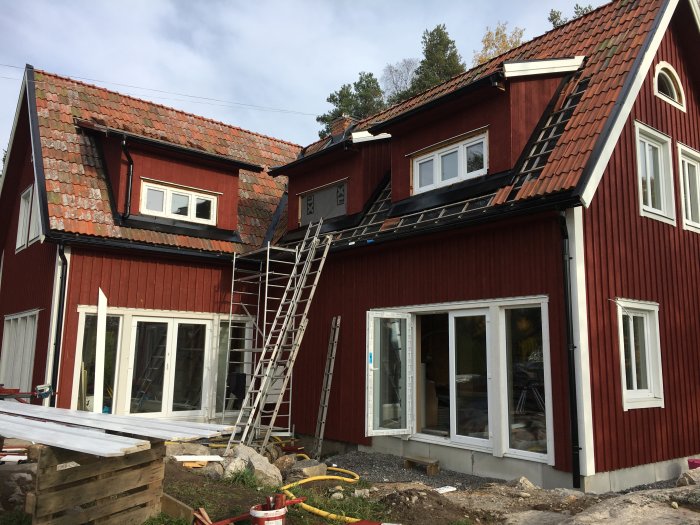 Rött hus under renovering med nyligen färdigställda takkupor, vindbrädor och takpannor, och en stege lutad mot fasaden.