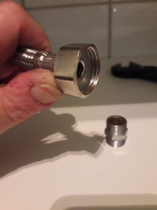 En hand håller en flexibel anslutningsslang bredvid en avtagen metallgängad adapter med olika gängdimensioner.