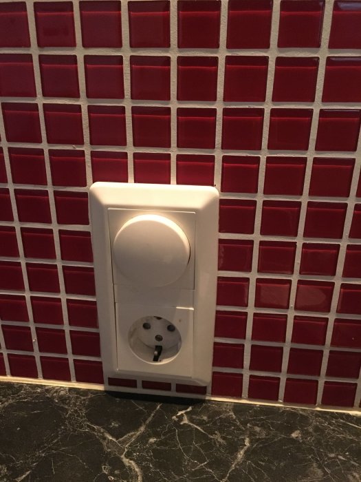 Infällt uttag med dimmer mot rödvit kakelvägg ovanför svart marmorbänk.